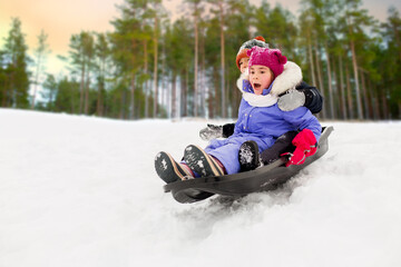 Fototapeta na wymiar childhood, sledging and season concept - group of happy little kids sliding on sled down snow hill in winter over snowy forest or park background
