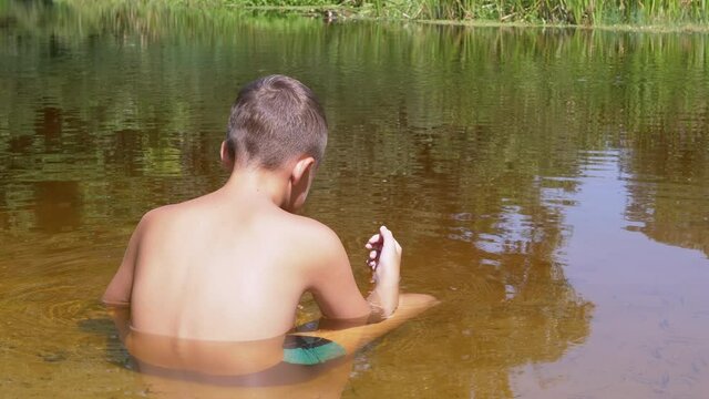 A Child Sits in a Lotus Position in a Shallow River at Sunset. Rear view of a teenager relaxing waist-deep in water. The boy watches the floating fishes in the lake in the background of green reeds.