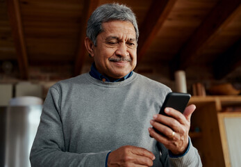 Smiling multi-cultural elderly male reading a message on smartphone while standing in modern kitchen.
