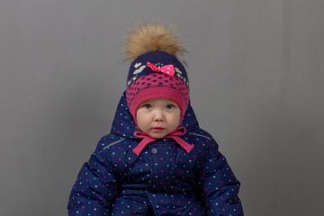 Cute baby girl 2 years old caucasian in warm winter clothes blue jumpsuit, hat, pink boots and mittens on a gray background