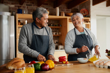 Multi-cultural elderly couple smiling, preparing meal in modern kitchen. Healthy retired lifestyle at home.