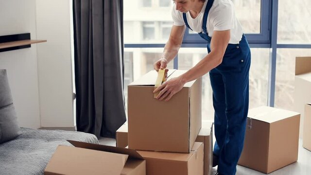 Packaging the box. Young male mover in blue uniform works indoors in the room