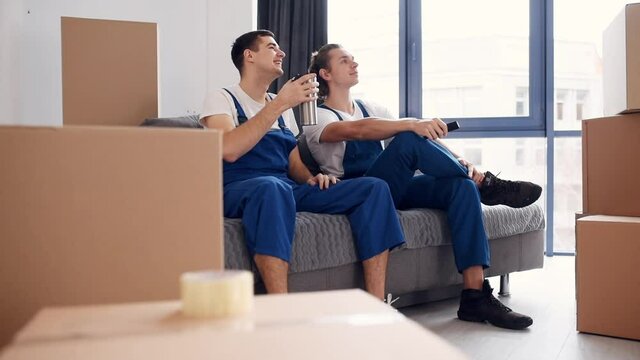 Tired and taking break. Two young movers in blue uniform working indoors in the room