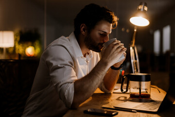 White bearded man drinking coffee while working with laptop in evening