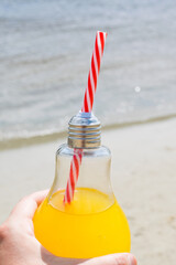 Fresh or cocktail in a glass bottle with a straw close-up in a female hand against the background of the beach and water, vertical format. Travel and recreation concept.
