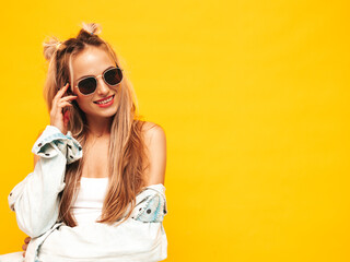 Fototapeta premium Portrait of young beautiful smiling blond female in trendy summer clothes. Sexy carefree woman posing near yellow wall in studio. Positive model having fun indoors. Cheerful and happy in sunglasses