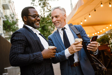 Multiracial two men using cellphone and talking while drinking coffee