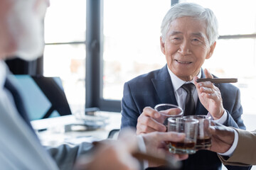 senior asian businessman with cigar clinking glasses with blurred multiethnic business partners in office