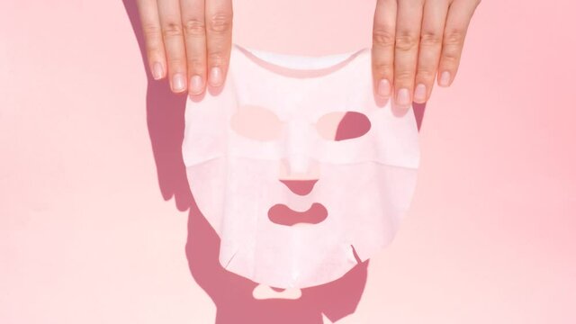 Female hands hold white sheet mask on pink background.
