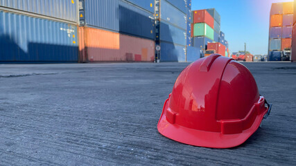 Red hard safety helmet hat for engineer or worker on concrete ground with container blurred image background