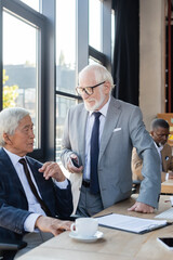 senior businessman with mobile phone talking to asian colleague near blurred contract on desk