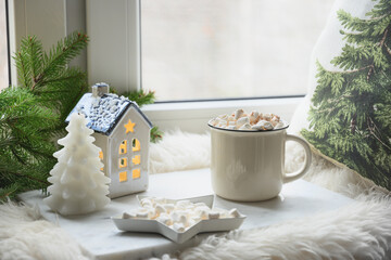 Winter coffee with marshmallow on windowsill with fir branches and little house as garland. Christmas cozy holiday season.