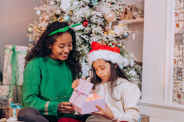 Fototapeta na wymiar Curly African American woman and brunette long-haired daughter wearing holiday hats open present box sitting against Christmas tree