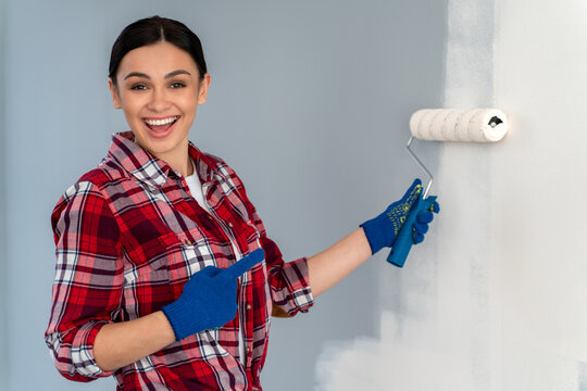 Overjoyed Young Caucasian Woman Pointing At The Roller At Her Hands While Enjoying Renovation Time At Home. Repair And Remodeling Concept