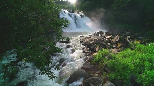 A powerful back lit waterfall in a deep forest in Kyushu Japan. Sekino falls. The river flows towards the camera under trees all in cinematic 4K slowmotion.