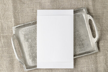 Blank white paper postcard on retro silver metallic tray on beige linen rustic table cloth. Empty greeting or invitation card mockup top view with copy space