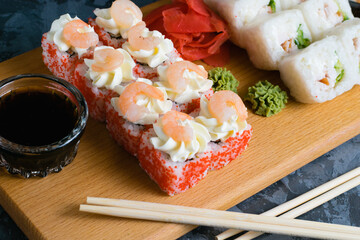 Appetizing and delicious sushi rolls with shrimp, cheese, caviar, chicken, soy sauce, nori, wasabi and ginger on a wooden board on a dark background
