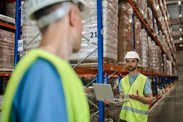 Man in overalls with laptop and colleague at warehouse
