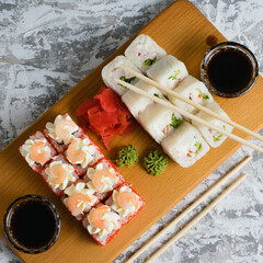 Delicious sushi rolls with chicken, cheese, shrimp, red ginger, wasabi and soy sauce on a wooden board on a light background with copy space