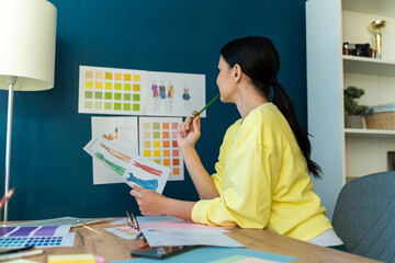Hardworking in office. Female fashion designer sitting at the table and examining sketches of her new collection while drawing at the workplace