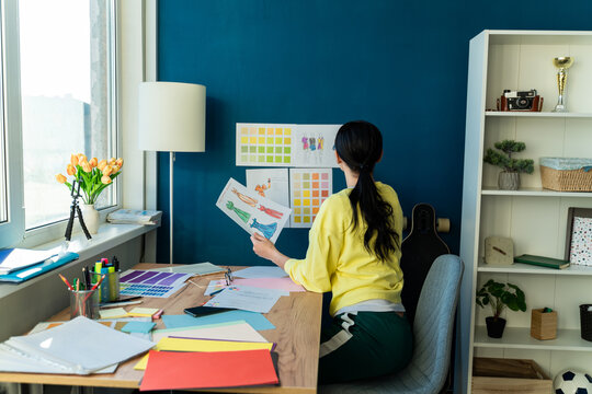 Back view of the young woman designer holding sketches of her new design collection while sitting at her apartments. Stock photo