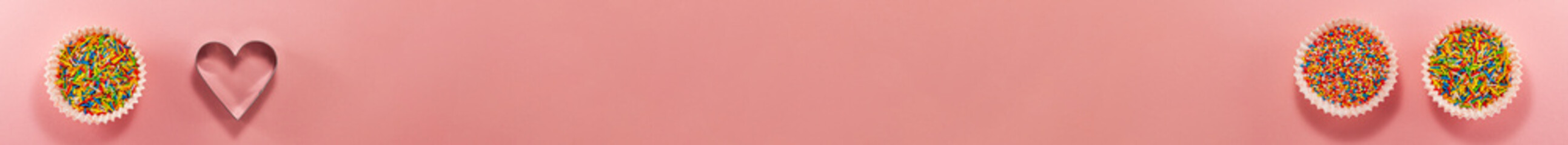 Banner of cupcake liners with colored sugar inside and heart shaped cutter on bottom from above. Flat lay top view. Confectionery concept with copy space on bright pink paper. National cooking day