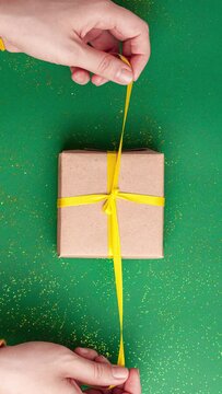 Human hands tied and untied brown gift box with a yellow satin ribbon bow on jade green background with gold colored glitter. Stop motion vertical animation Christmas Holidays and present flat lay