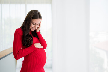 Fototapeta premium Pictures of new mothers, Europeans, Russians, pregnant Standing by the window, holding his tummy, smiling happily in the bedroom. wearing red maternity clothes