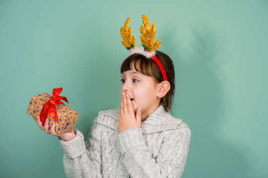 Studio portrait of suprised child girl wearing yellow deer horns with gift box in hand on mint background