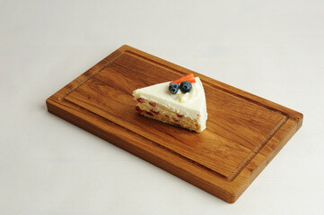 Piece of milk cake on wooden board. Biscuit layers, butter cream, berry jam, cheese glaze. Decorated with fresh berries