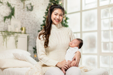 Beautiful Asian woman holding newborn baby in arms sit on bed at white cozy home with christmas tree in background.Happy infant baby sleep in mother arms safety and comfortable.Mom and Baby Concept