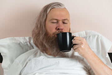 Bearded man enjoying his morning coffee in bed to cheer up before starting new day. Closeup.