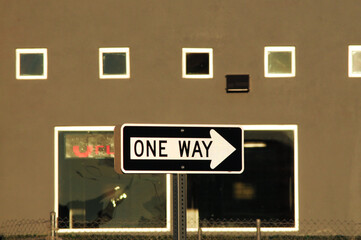 A one-way traffic sign is positioned in front of a commercial building facade with square windows...