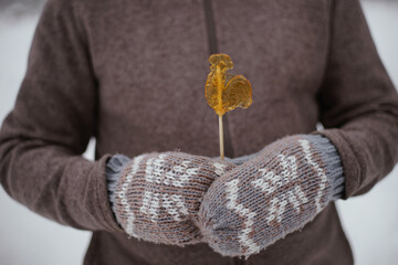 Caramel on a stick figure of a Cockerel in his hands in mittens.