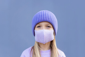 Cute little caucasian girl eight years old with blonde hair and protective face mask smiling outdoor. Kid wearing stylish shirt and knitted hat violet color. Trendy color of the 2022 year very peri