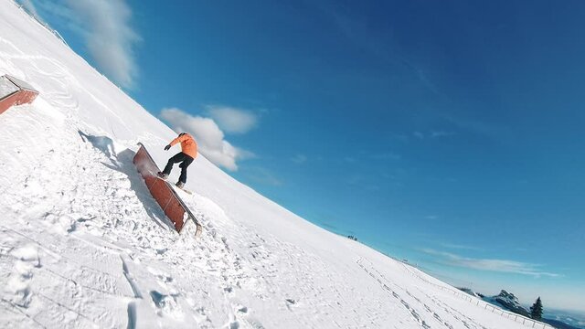 Snowboarder with shining orange suit filmed by fpv drone while doing tricks on the rail bank 60fps