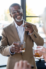 senior african american businessman with cigar and glass of whiskey smiling near blurred colleague in office