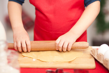 Kid boy or girl in red apron baking Christmas cookies at home. Child rolls the dough onto cookies.