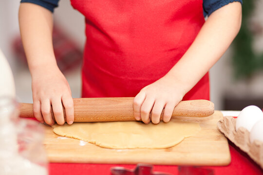 Kid Boy Or Girl In Red Apron Baking Christmas Cookies At Home. Child Rolls The Dough Onto Cookies.