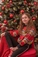 Teenage girl in a New Year's sweater near the Christmas tree. Girl 11 years old on a red sofa.