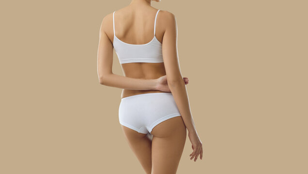 Cropped shot of young female model in underwear. Back view of beautiful Caucasian woman with fit, attractive body wearing basic soft white bra and underpants posing isolated on solid beige background