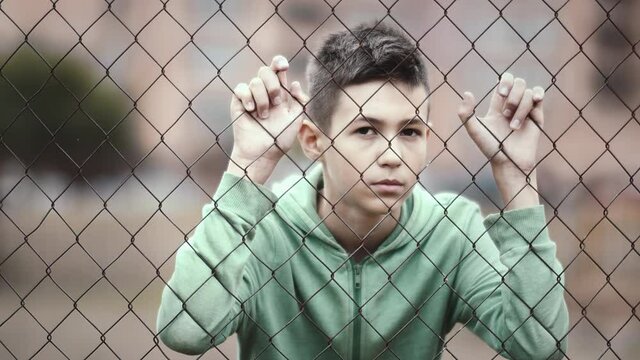 refugee boy stands alone head bowed near the fence, frustrated boy dropped eyes, regrets the act, cinematic shot
