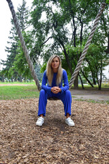 Fototapeta premium Attractive blonde woman sitting on a hanging rope swing in a blue suit in the park
