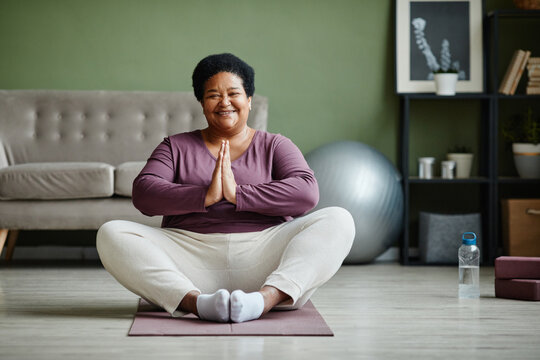 Full length portrait of senior black woman sitting on floor in lotus position and smiling at camera while enjoying yoga at home, copy space