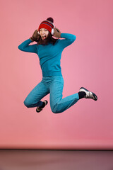 Full-length portrait of excited young girl in warm winter blue sport attire jumping isolated on pink studio background. Concept of emotions