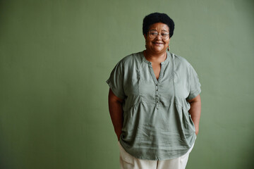 Waist up portrait of black senior woman smiling at camera while standing against green background,...