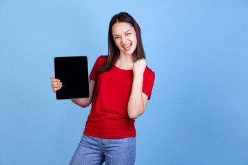 Emotional young beautiful girl in red t-shirt using digital tablet isolated on blue studio...