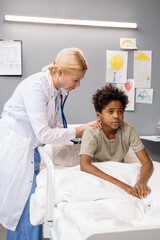 Obraz premium Mature female doctor in white coat examining the little boy with stethoscope while he sitting in bed at hospital ward, she listening to his breathing