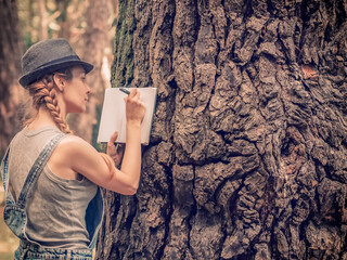 A fair-haired girl in a hat makes notes in a notebook leaning on a massive trunk of a forest tree