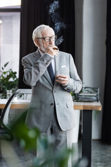 senior businessman in suit smoking cigar while holding glass of whiskey